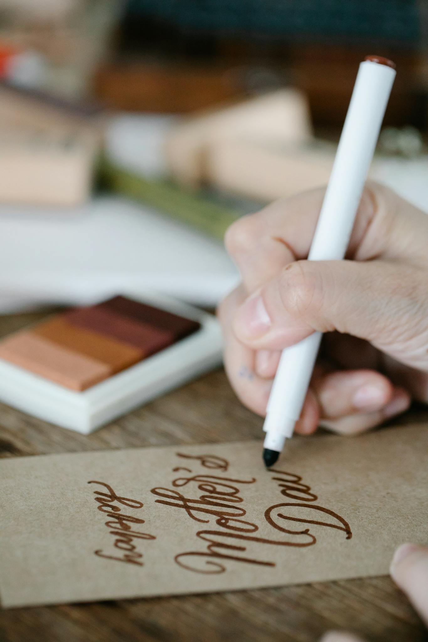 Close-up of hand crafting a Mother's Day card with calligraphy, pen, and art materials.